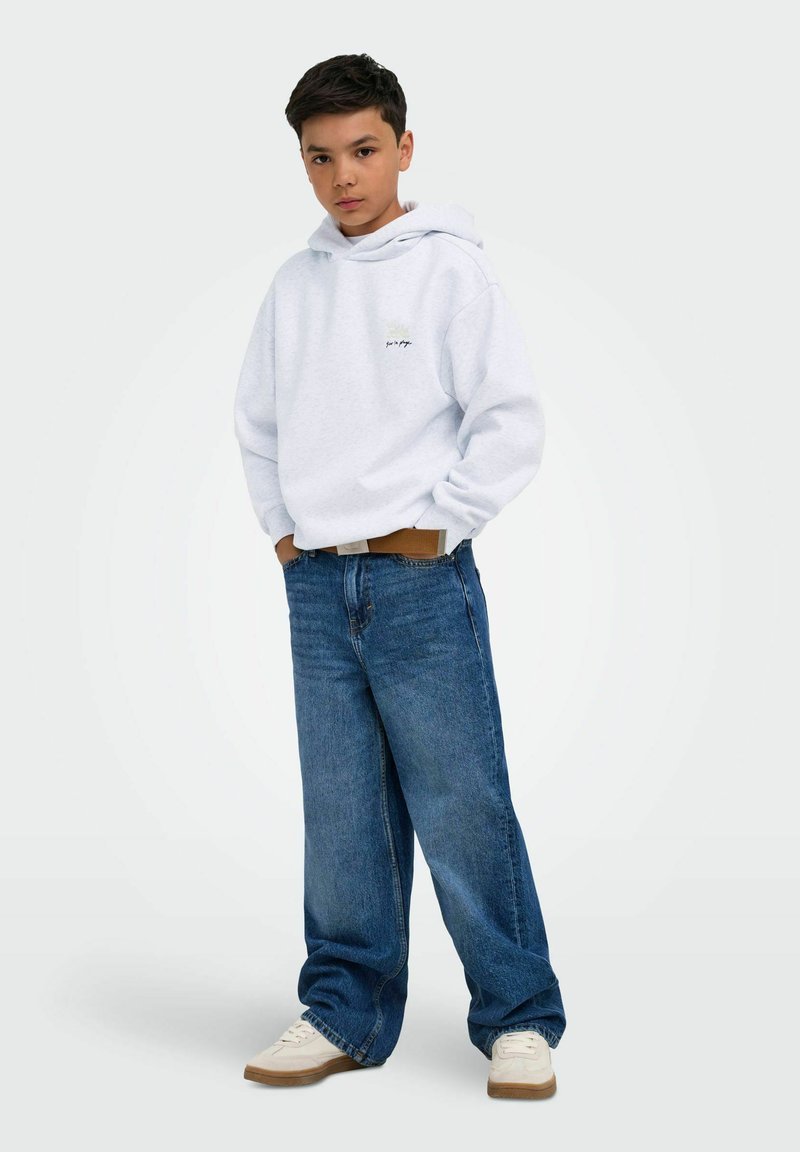 Garçon debout portant un sweat à capuche blanc, un jean bleu ample, des baskets beiges, les mains dans les poches sur un fond clair.