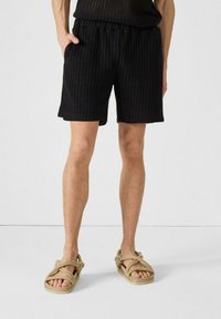 Pantalones cortos de malla negra con cintura elástica, que incluyen bolsillos laterales y un corte cómodo. Combinados con sandalias beige anudadas.