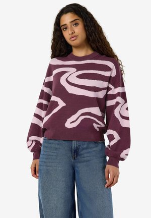 Femme aux longs cheveux bouclés portant un pull grenat avec des motifs tourbillonnants rose clair et un jean bleu sur un fond uni.