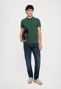 Polo de manga corta verde con cuello y logo bordado, combinado con jeans azul oscuro y mocasines beige claros, sosteniendo una chaqueta azul marino.