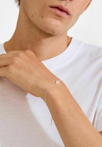 Zilveren armband met een delicate ketting en een bloemmotief, gedragen om een pols. De achtergrond is licht, met een wit T-shirt zichtbaar.