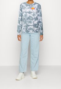 Sudadera azul claro con estampado tie-dye y logo gráfico blanco, combinada con jeans azul claro de corte holgado y zapatillas blancas.