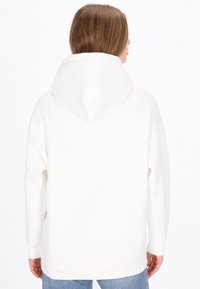 Sudadera blanca con capucha, de ajuste holgado, con aberturas laterales y puños acanalados. Textura suave y diseño minimalista. Combinada con vaqueros azules.