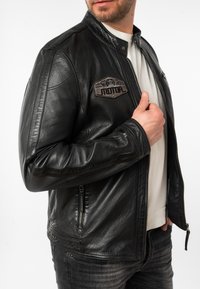 Zwarte leren motorjack met een hoge kraag, ritssluiting aan de voorkant, geribbelde schouderaccenten en een decoratieve patch op de borst.
