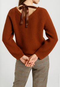 Maglione a coste color ruggine con scollatura a V e un nastro di raso legato sul retro. Il modello indossa pantaloni a quadri.