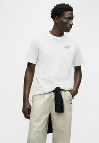 Mann som ser til høyre, iført hvit kortermet skjorte med liten logo på brystet og lyse beige bukser med sort plagg knyttet rundt livet.