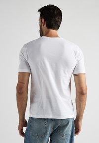 Vit bomullströja med korta ärmar, rund halsringning; bärs med blå jeans. Synlig tatuering på armen som säger "NO MERCY."