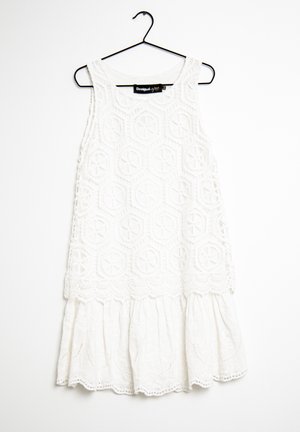 Biała sukienka bez rękawów z okrągłymi wzorami koronkowymi i falistym dołem, zawieszona na czarnym metalowym wieszaku na białym tle.
