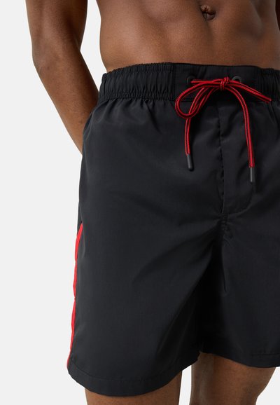 Homme portant un short de bain noir avec des bandes latérales rouges et un cordon rouge, montrant le torse et le haut des jambes sur un fond uni.