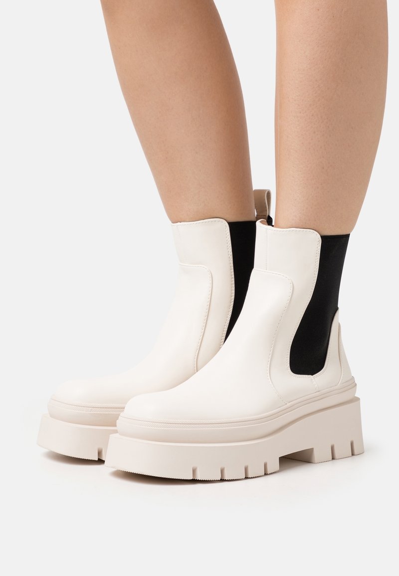 Bottes Chelsea blanches à plateforme avec semelle épaisse côtelée et panneaux latéraux élastiques noirs, portées sur jambes nues sur fond blanc.