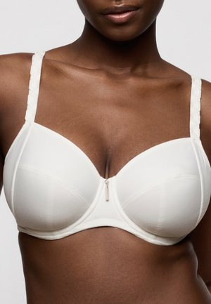 Close-up van een donkerharige vrouw die een witte beugelbeha draagt met decoratieve bandjes en een klein hangertje in het midden.