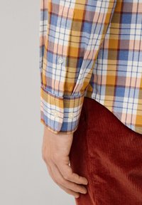 Camisa de cuadros en tonos amarillos, azules y rosas, con mangas largas y puños con botones. Usada sobre pantalones de pana rojos.