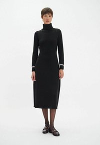 Robe noire à col montant avec des manches longues, coupe slim, longueur genoux, ornée d'accents blancs aux poignets. Le mannequin porte des chaussures sombres et des collants transparents.