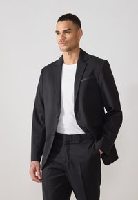 Jachetă neagră de costum cu două butoane și rever în formă de V, combinată cu un tricou alb. Conține un buzunar pe piept și are o croială elegantă, ajustată.