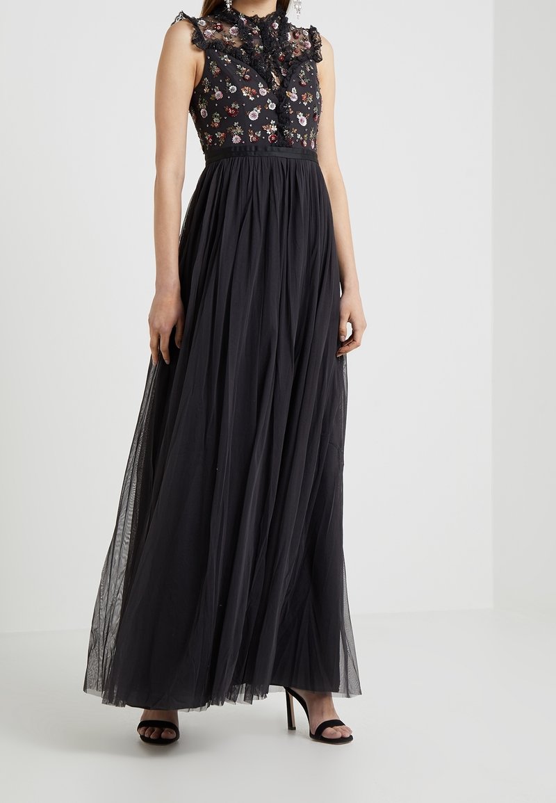Robe maxi noire avec corsage brodé de fleurs, col à volants en tulle transparent, taille cintrée et jupe plissée en tulle ; associée à des escarpins noirs.