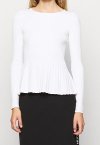 Weißes, geripptes Langarmoberteil mit Peplum-Taille, Rundhalsausschnitt und strukturiertem Finish, kombiniert mit einem schwarzen Rock, der ein Logo-Element zeigt.