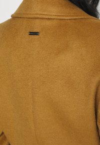 Brun ullkappa med mjuk textur, utrustad med krage och en märkesskylt bak. Materialet har en slät finish.