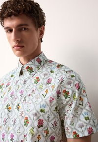 Chemise à manches courtes avec une base blanche ornée d'un motif floral coloré, petit col, et un design texturé subtil.