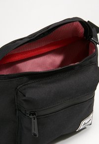 Černá plátěná taška s červeně pruhovanou podšívkou. Obsahuje přední kapsu na zip a hlavní prostor se zipovým uzávěrem v černé barvě.