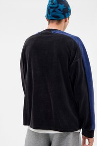 Sweatshirt oversized em cordura preta com painéis de ombro texturizados em azul. Tecido suave, textura rib elevada e ajuste descontraído. Usado com calças cinzentas.
