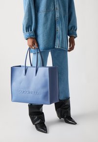 Sac tote Coccinelle, en cuir texturé bleu clair, dispose de deux poignées robustes et d'un logo en relief à l'avant.