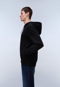 Schwarzer Kapuzenpullover mit Kordelzug, Kängurutaschen und gerippten Bündchen; kombiniert mit blauen Jeans, dezentes Branding auf der Brust.