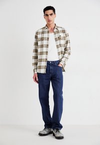 Noor mees seisab käsi taskus, seljas ruuduline särk valge tanki üle, sinised teksad ja heledate laced-sussidega, valge tausta ees.