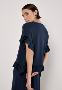 Marineblaue Bluse mit kurzen, ausgestellt geschnittenen Ärmeln und Rüschensaum. Weicher, strukturierter Stoff mit lockerer Passform, der eine elegante, fließende Silhouette bietet.