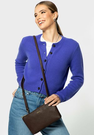 Mujer sonriendo con un cárdigan azul abotonado, vaqueros azul claro y llevando un bolso bandolera marrón oscuro sobre el hombro.