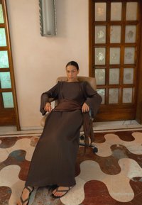 Ruskea, laskostettu maksimekko pitkillä hihoilla ja vyötärösoljella, yhdistetty mustiin flip-floppiin, istuen tuolilla puuoven edessä.