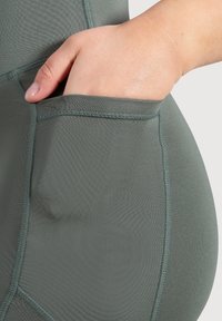 Ciemnozielone legginsy sportowe z boczną kieszenią. Materiał jest gładki i elastyczny, z subtelnymi przeszyciami wzdłuż szwów.
