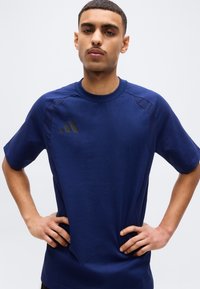 Νέος άνδρας με κοντά μαλλιά, φορώντας μπλε ναυτικό αθλητικό μπλουζάκι με μαύρο λογότυπο Adidas, στέκεται με τα χέρια στη μέση μπροστά σε έναν απλό φόντο.