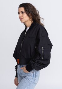 Black bomber jacket with a cropped design, zip front, ribbed cuffs, two zippered side pockets, and an orange label at the hem.