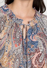 Blus med paisleymönster i blått, orange och grått. Har en rynkad halsringning och en nyckelhålsdetalj med knytband.