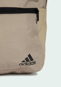 Zaino in tessuto beige con superficie testurizzata, cerniera nera e logo Adidas nero stampato sul davanti.