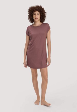 Frau mit lockigem Haar, barfuß, trägt ein mauvefarbenes Kleid mit kurzen Ärmeln, das über das Knie reicht, vor einem schlichten, hellen Hintergrund.