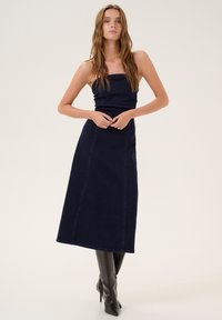 Dark denim maxi dress with a fitted bodice and straight skirt, featuring contrast stitching and a minimalist design. Paired with knee-high boots.