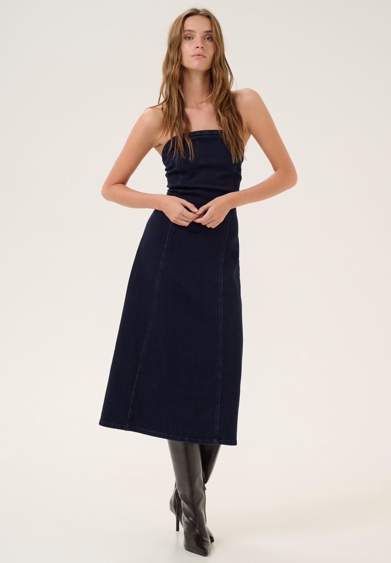 Dark denim maxi dress with a fitted bodice and straight skirt, featuring contrast stitching and a minimalist design. Paired with knee-high boots.