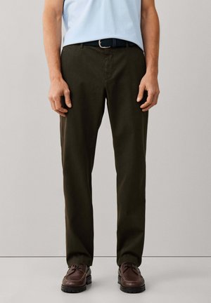 Hombre con pantalones rectos de color verde oscuro, camisa azul claro, cinturón negro y zapatos de cuero marrón oscuro, de pie frente a un fondo liso.