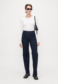 Tummansiniset korkeavyötäröiset farkut, jotka on valmistettu denimistä, yhdistettynä istuvaan valkoiseen pitkähihaiseen puseroon ja mustiin asusteisiin, kuten aurinkolaseihin ja laukkuun.