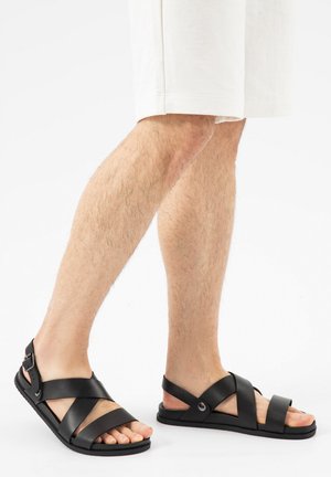 Benen met witte korte broek en zwarte leren sandalen met gekruiste bandjes en gesp, staand op een witte achtergrond.