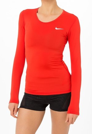 Kobieta w dopasowanej, czerwonej koszulce Nike z długim rękawem i czarnych szortach sportowych, pozująca na białym tle.