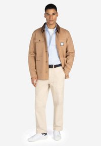 Chaqueta utilitaria marrón con cuatro bolsillos, cierre de botones y un parche. Combinada con una camisa azul clara, pantalones crema y zapatillas blancas.