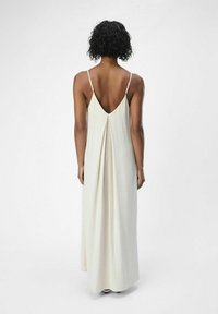 Abito maxi color crema chiaro, realizzato in un tessuto testurizzato. Presenta uno scollo a V profondo sul retro e spalline sottili e regolabili, con una silhouette fluida.