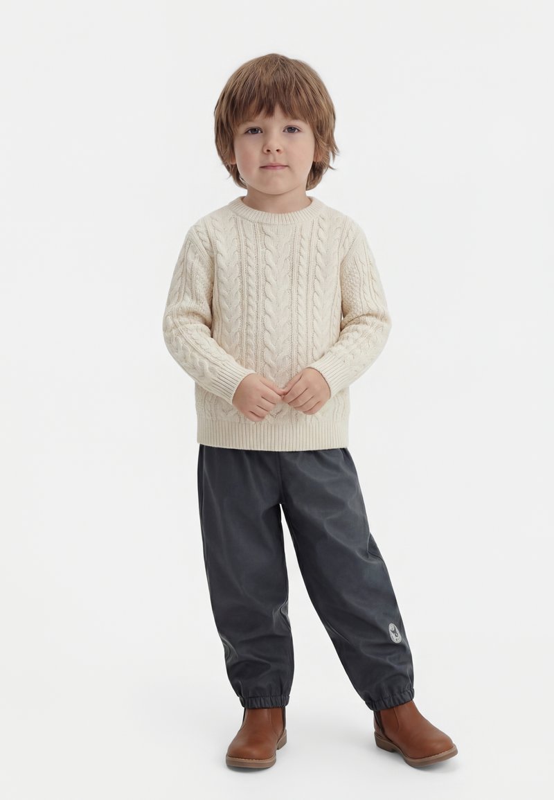 Jeune enfant aux cheveux bruns portant un pull en maille torsadée crème, un pantalon foncé à poignet élastique et des bottines marron, debout de façon neutre sur fond blanc.