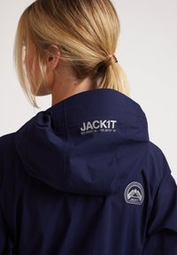 Chaqueta impermeable azul marino con capucha, con el logo gris "JACK1T" y coordenadas geográficas en la parte posterior, textura suave y diseño minimalista.