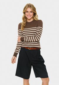 Donna sorridente che indossa un maglione a righe marrone e beige, pantaloni corti neri fino al ginocchio e una cintura marrone su uno sfondo bianco semplice.