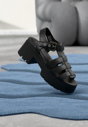 Black chunky platform sandal with buckle strap, displayed above a textured blue surface and blurred background.