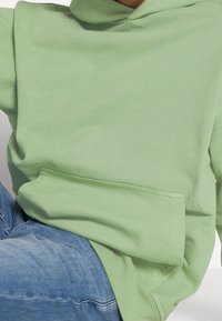 Sudadera verde claro con capucha, confeccionada en algodón suave, con un bolsillo frontal y un detalle sutil de logo en el dobladillo. Combinada con jeans azules.