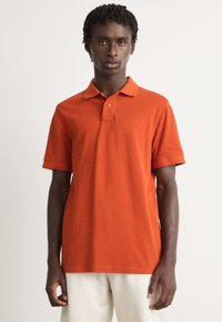 Ung mann med korte dreadlocks iført en oransje polo-skjorte og lyse bukser, stående mot en ensfarget hvit bakgrunn.